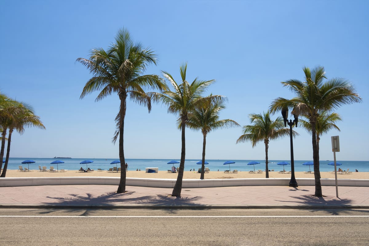 Las Olas Beach in Fort Lauderdale, Florida, with palm trees lining the walkway and blue umbrellas dotting the sandy shore