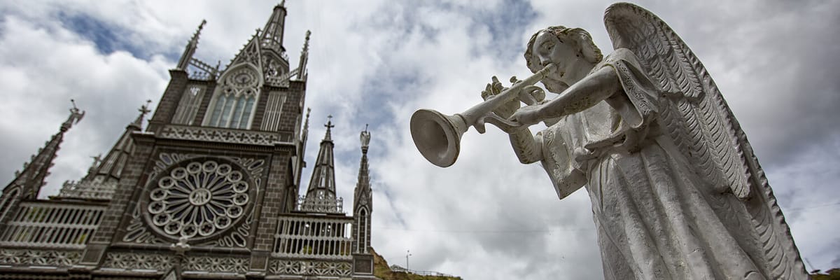 Primer plano de una estatua de รกngel con trompeta frente al Santuario de Las Lajas en Ipiales, Colombia. Descubre este icรณnico destino religioso y arquitectรณnico con vuelos a Ipiales, la 'Ciudad de las Nubes Verdes