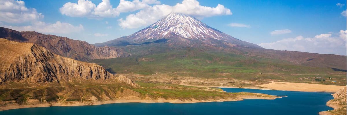 Snow‑topped volcanic peak of Mount Damavand in Iran rising above a green‑brown plateau and a shimmering lake.