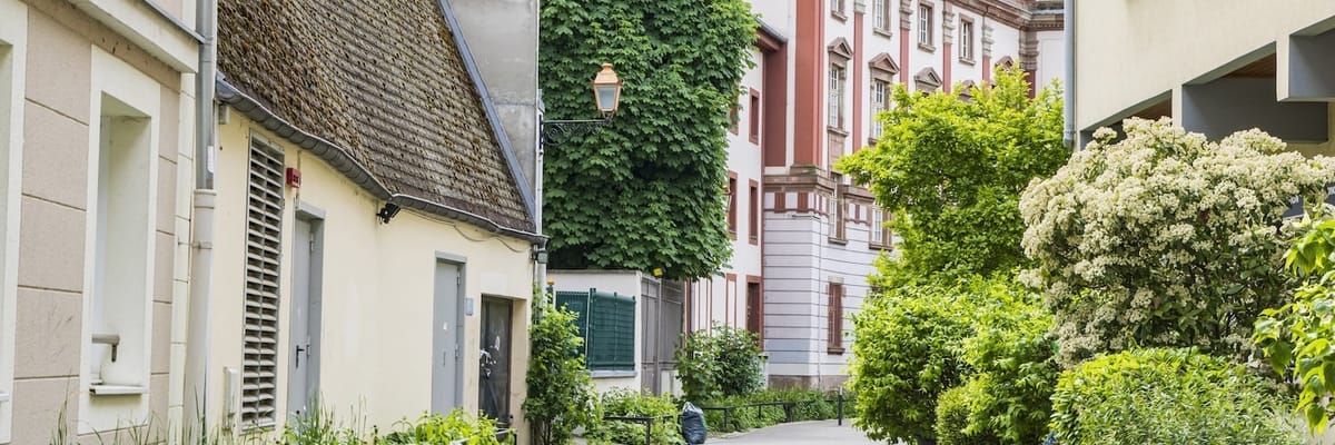 Narrow cobbled path lined with flowering shrubs and pastel buildings in a peaceful residential neighborhood.