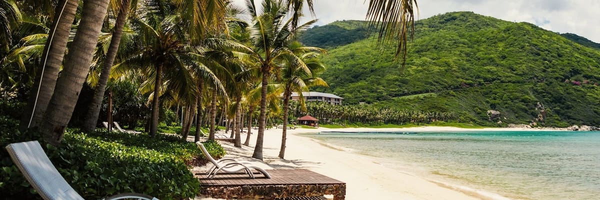 Empty lounge chairs under palm trees line a secluded tropical beach with lush green hills in the background.