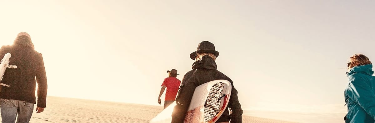 Group of surfers walking toward the ocean across sunlit sand dunes, boards in hand, under a soft sky.