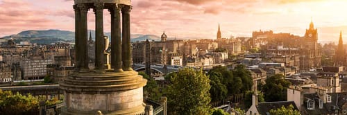 View of Edinburgh, Scotland, featuring the iconic Dugald Stewart Monument in the foreground and Edinburgh Castle prominently in the background, set against a vibrant sunset sky