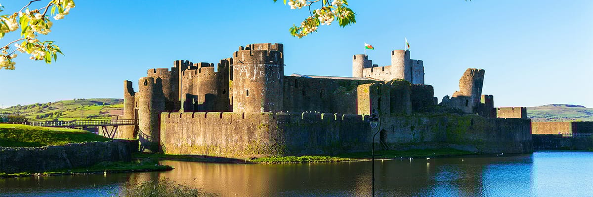 A castle in Cardiff surrounded by a lake and green hills with blue sky in the background.