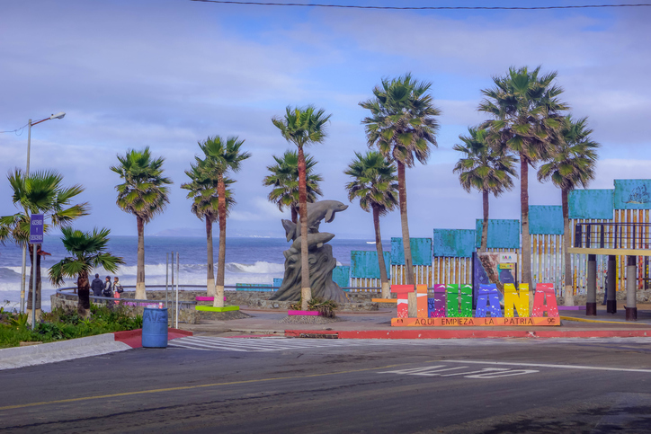 Cartel de Tijuana en plazoleta con palmeras frente a la playa, cerca de vallas del muro fronterizo.