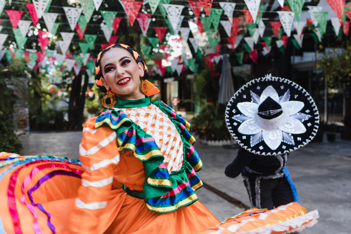 A latin couple dressed in colonial mexican suits dancing in the street, with mexican flags in the background.