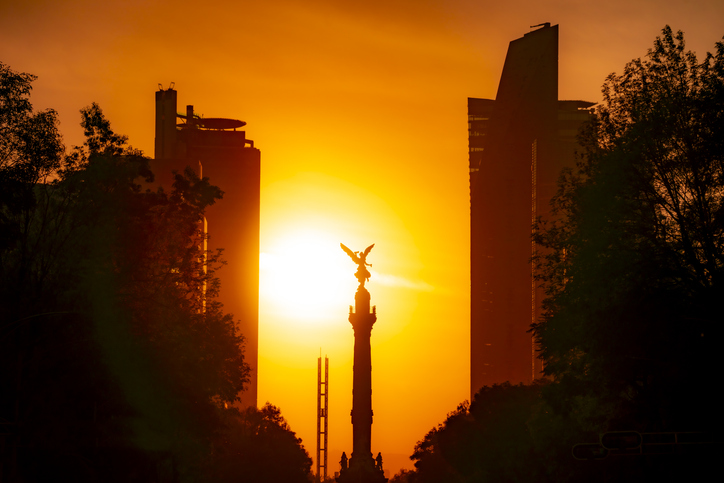 The Angel of Independence statue in Mexico city between tall buildings against a colorful orange sunset sky.