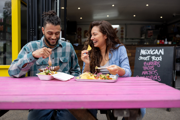 A young couple sitting on a pink table and enjoying Mexican street food.