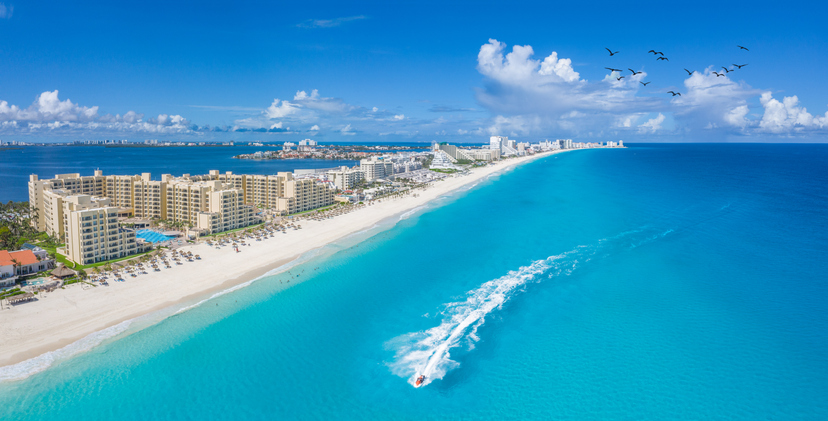 Vista aérea de la zona hotelera de Cancún, con hoteles frente al mar y una lancha cruzando el Caribe.