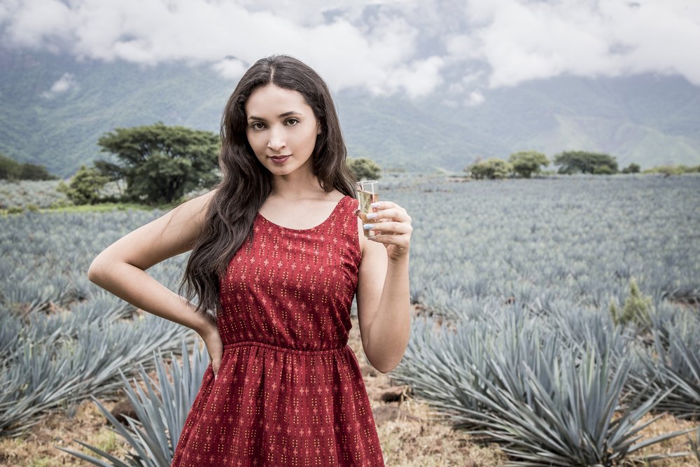 Una mujer joven sostiene un shot de tequila frente a las plantaciones de tequila.