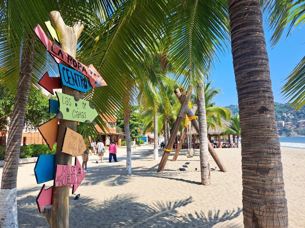 Señalización artesanal en la playa de Ixtapa, con indicaciones hacia lugares como Playa Las Gatas y el Museo Arqueológico.