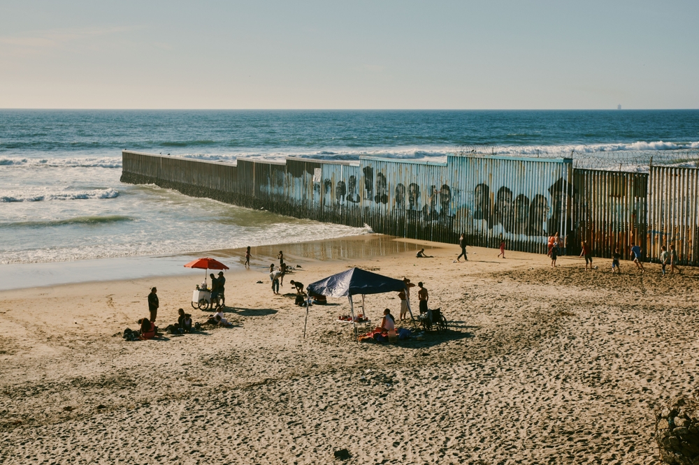 Vista panorámica del muro fronterizo en Playas de Tijuana, con gente disfrutando del mar bajo sombrillas.
