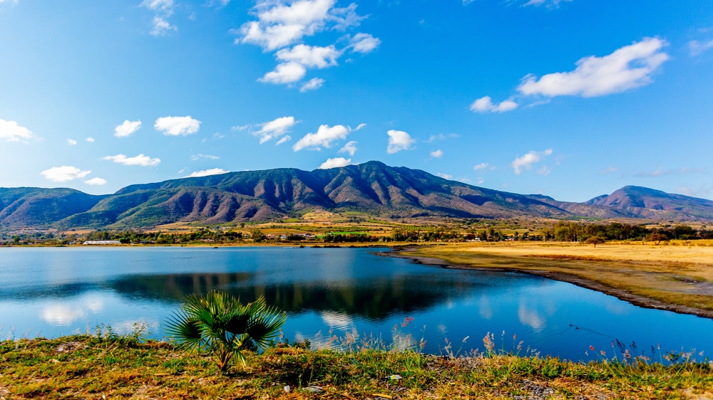 Panoramic Mexican mountain landscape, lake with mirror-like reflection, surrounded by arid land and farms, sunny day along the Guadalajara to Chapala highway.