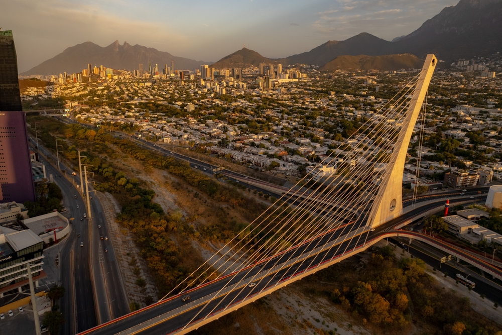 Vista aérea panorámica de la ciudad de Monterrey al atardecer, con un puente autopista al frente y el Cerro de la Silla al fondo.