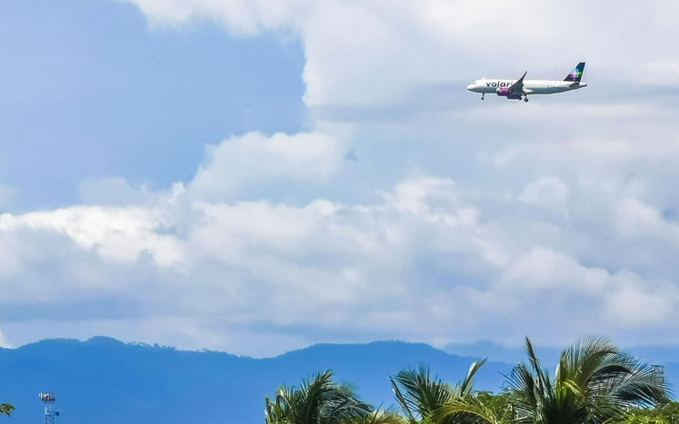 Volaris airbus flying low across a cloudy sky, above mountains and palm trees.