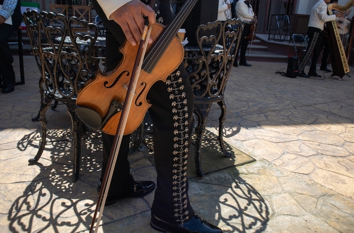 Músico vestido con clásico pantalón mariachi sosteniendo un violín junto a las mesas de un bar.