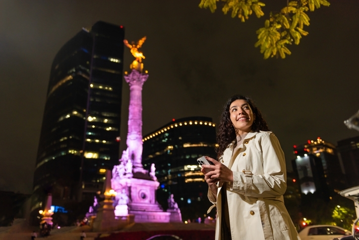 Mujer casual sostiene su celular frente a la estatua del Ángel de la Independencia por la noche.