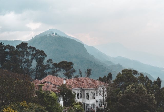 Casa colonial con tejado rojo rodeada de vegetación andina en las montañas de Bogotá, Colombia, con Monserrate al fondo entre la neblina.