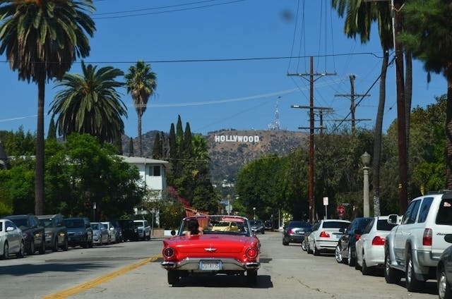 Vista desde un automóvil clásico rojo en una calle de Los Ángeles, con palmeras y el cartel de Hollywood visible al fondo.
