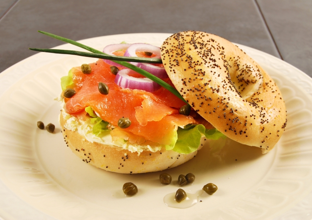 Bagel abierto con queso crema, salmón ahumado, alcaparras, lechuga, rodajas de cebolla morada y cebollín, servido en un plato blanco.