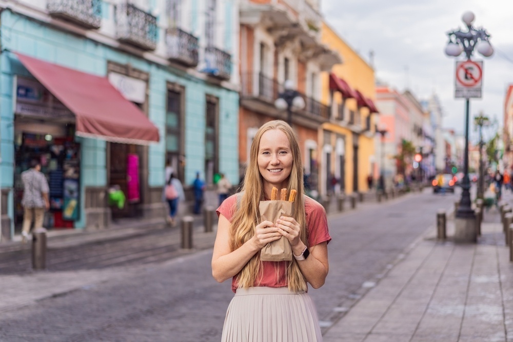 Turista comiendo churros en el centro histórico de Puebla, con coloridos edificios de fondo.