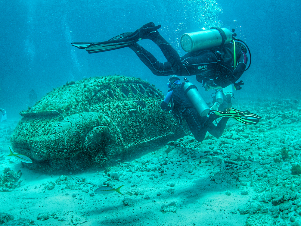 Dos personas equipadas con trajes de buceo y tanques de respiración admiran un coche en las profundidades del mar, en MUSA, Cancún.