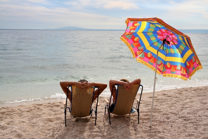 Two people sitting by the seashore take a rest next to a beach umbrella.