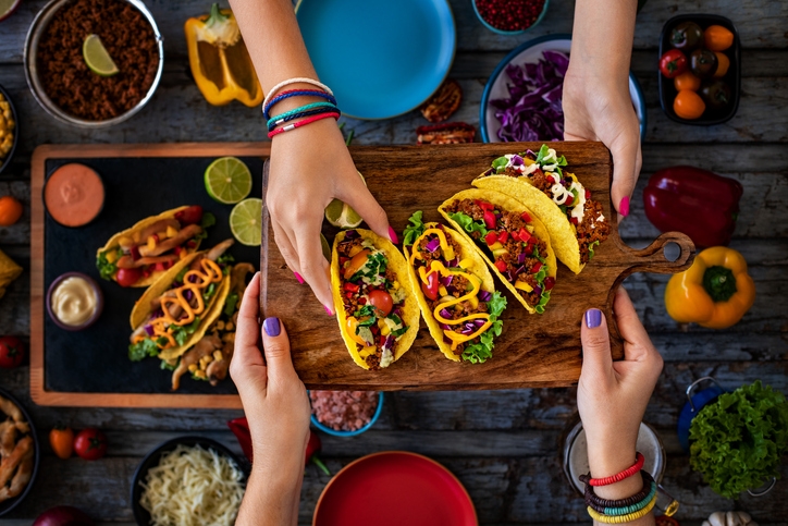 The hands of two people holding a wooden plate with a variety of Mexican tacos, above a served table full of tacos and typical Mexican dressings.
