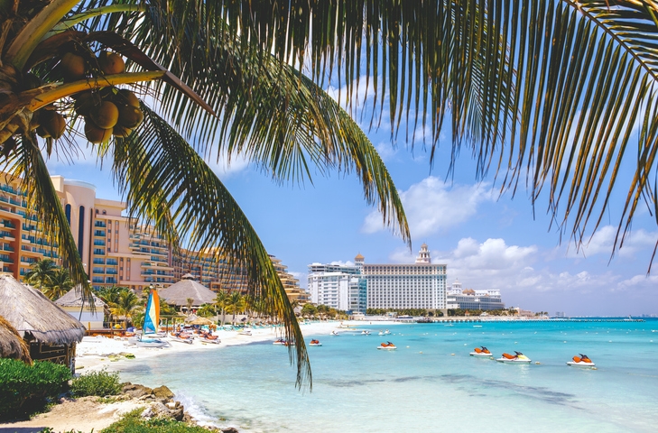 Playa en Cancún con palmeras, aguas turquesa y hoteles al fondo.